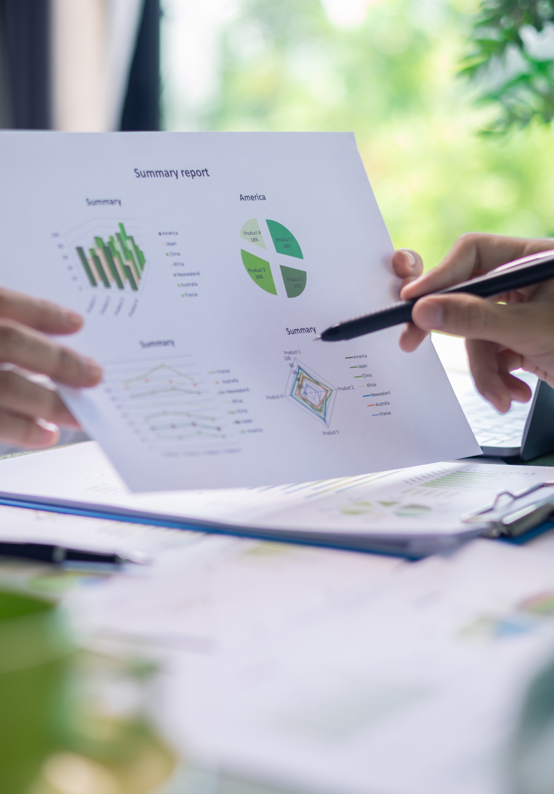 Due persone in abbigliamento professionale analizzano un report stampato con grafici e diagrammi, sedute a un tavolo circondato da oggetti verdi e materiale d’ufficio, in un ambiente luminoso e naturale.