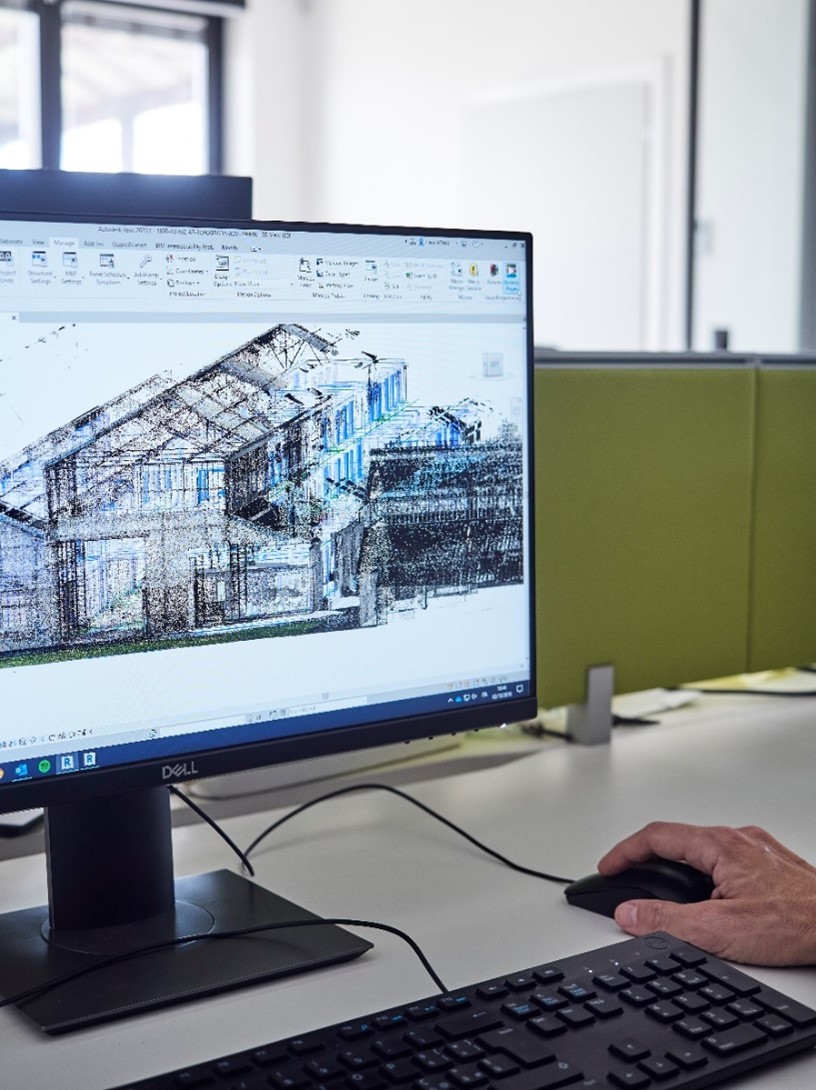 Vista ravvicinata di uno schermo su cui è visualizzato un modello 3D architettonico in Autodesk ReCap, con una mano che utilizza il mouse per navigare il progetto digitale in un ufficio tecnico moderno.