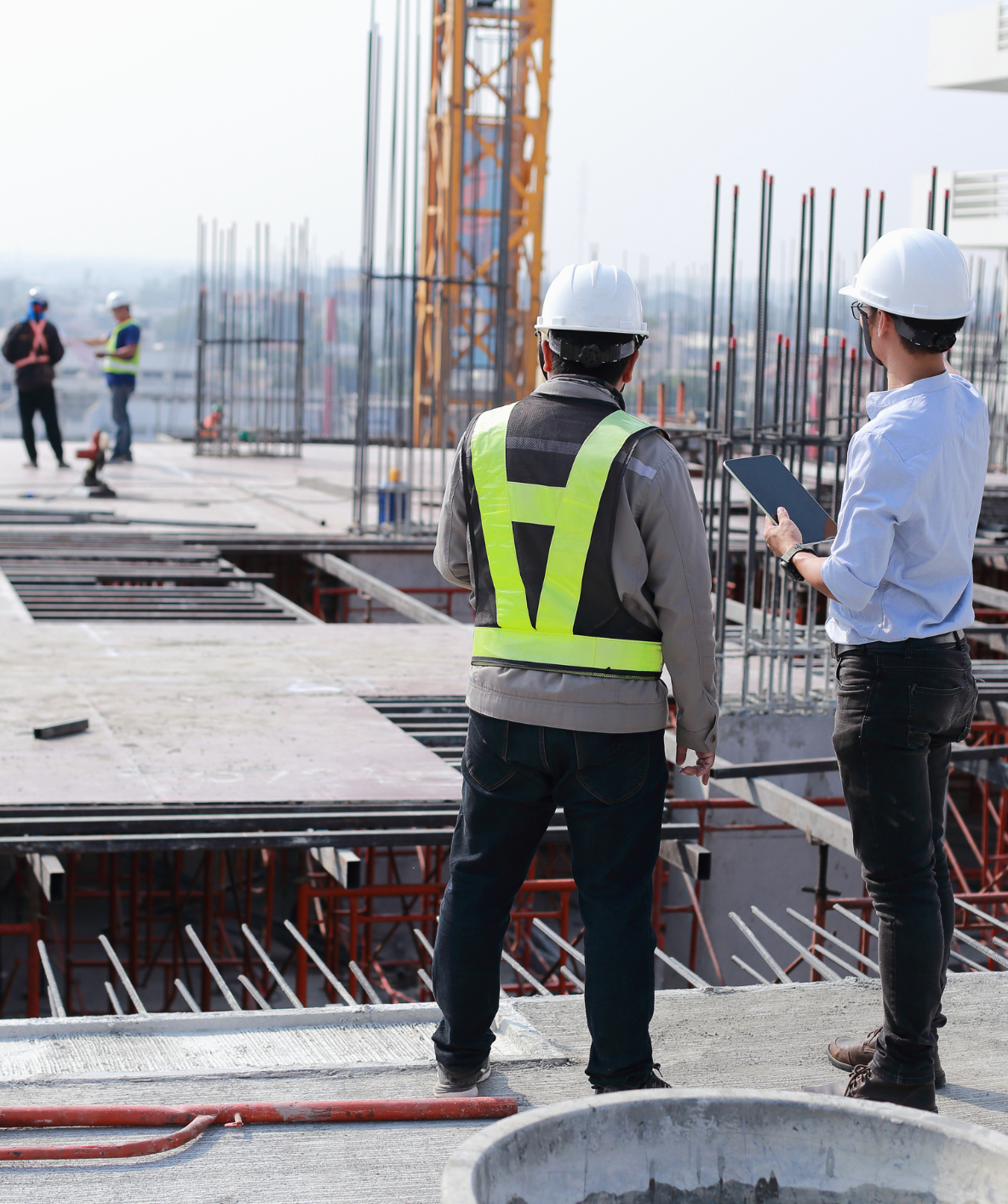 Operai e tecnici in cantiere osservano l'avanzamento di una costruzione in calcestruzzo. Indossano caschi e giubbotti di sicurezza.