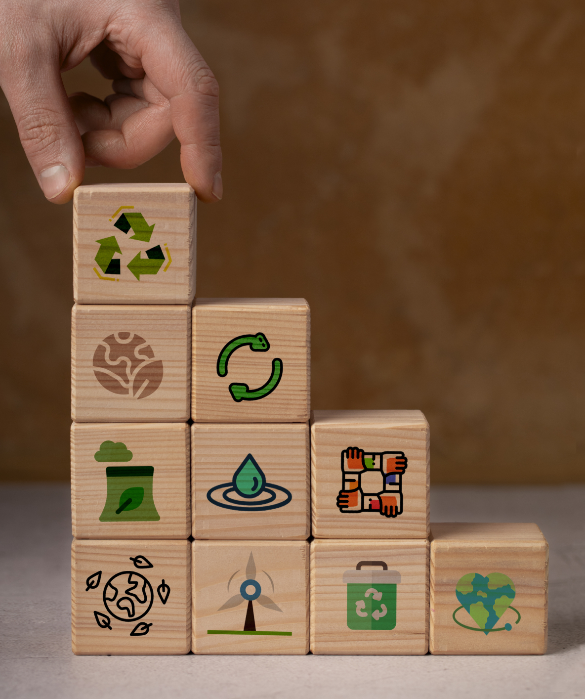 Una mano posiziona un cubo di legno con il simbolo del riciclo su una piramide di blocchi illustrati con icone legate alla sostenibilità: energie rinnovabili, economia circolare, acqua, comunità, e cura del pianeta.