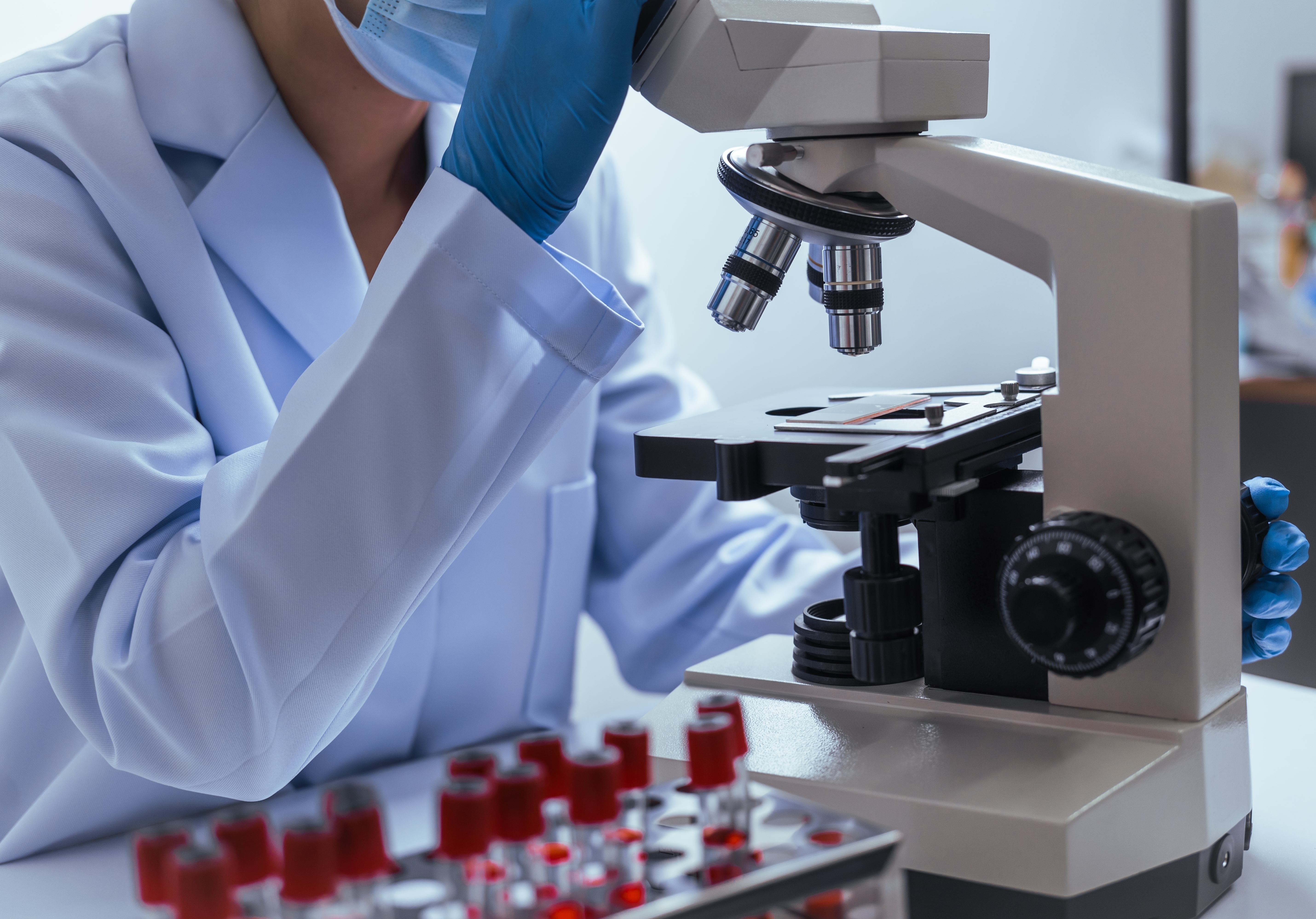 Mani di ricercatrice a lavoro in un laboratorio scientifico moderno con microscopio circondato da provette e strumenti di analisi.